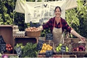 Farmers market vendor behind a stall displaying peaches, tomatoes, lemons, and leafy greens.