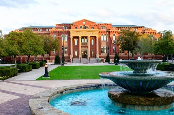 A large, two-story brick building with a clock tower, framed by trees, a front lawn, and a fountain in Southlake Town Square.