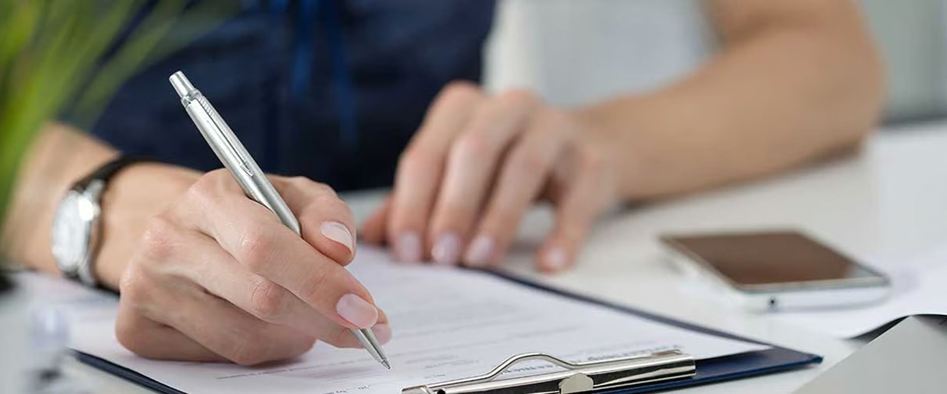 A person signs paperwork with a silver pen beside a smartphone.
