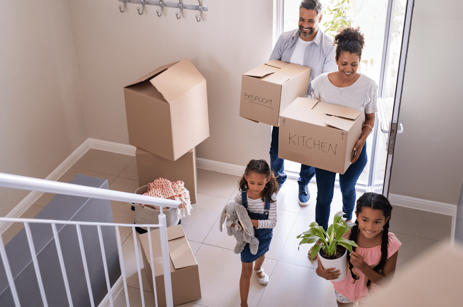 Parents carry moving boxes as their two daughters bring a stuffed animal and potted plant into the home.