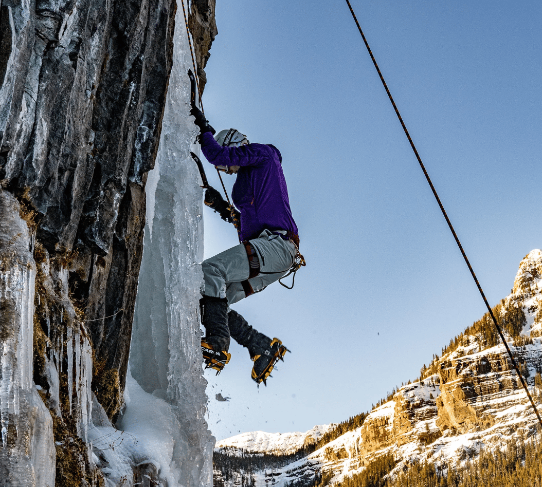 Ice climber scaling a frozen cliff above snow-covered mountain slopes.