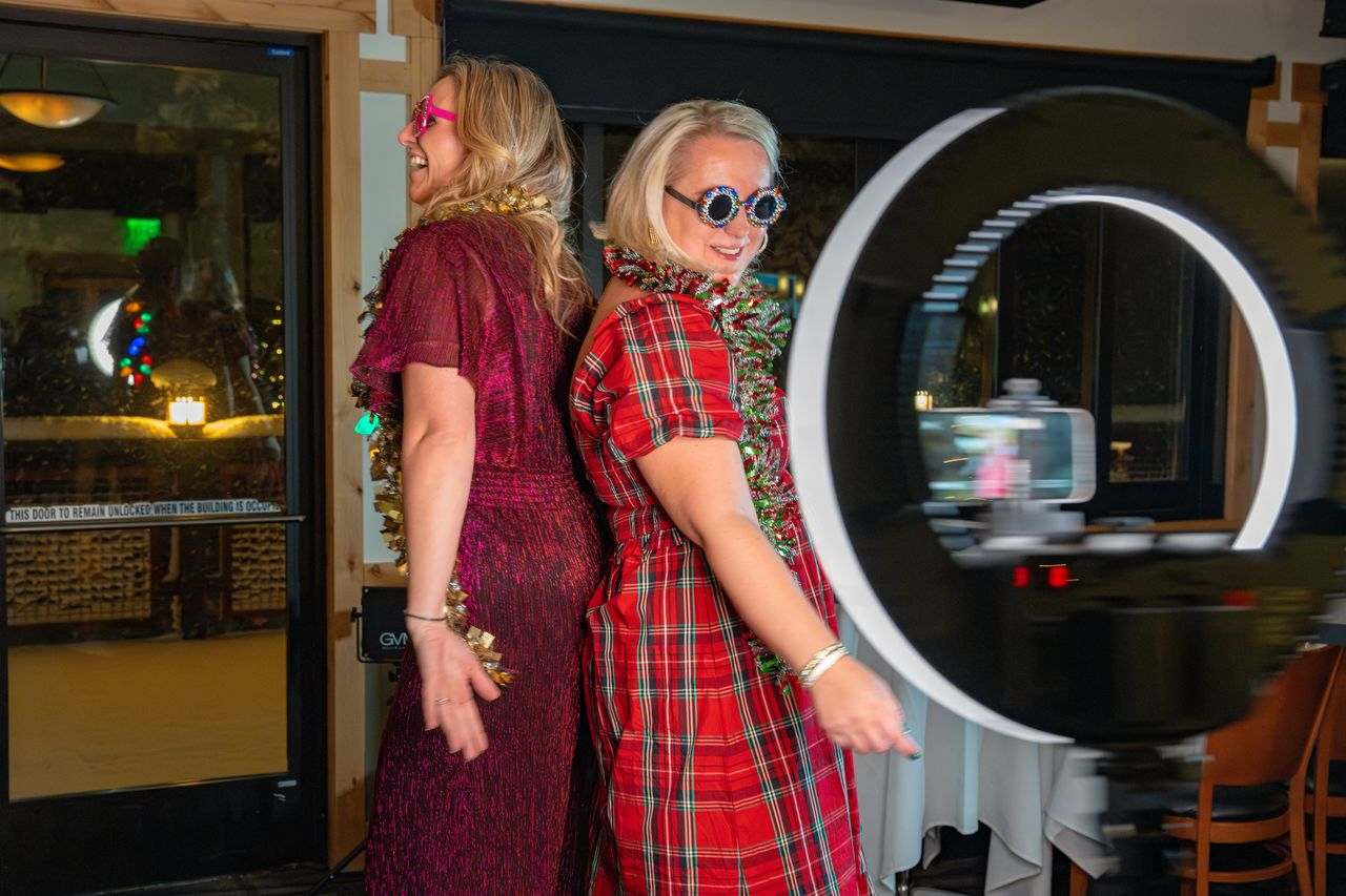 Two women pose back-to-back in festive outfits beside a circular photo booth light.