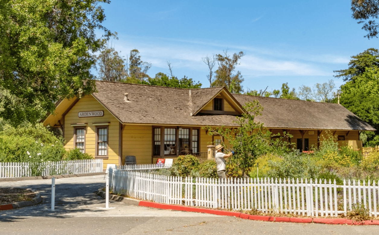 The image shows a yellow house with a white picket fence in front of it. 