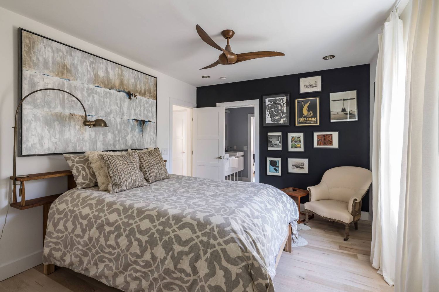 Bedroom with a patterned bed, dark gallery wall, cream armchair, and wood ceiling fan.