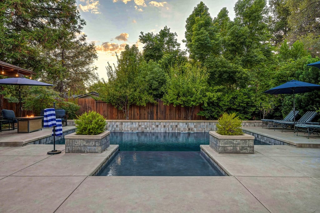 Backyard pool with a shallow tanning shelf, raised planters, and patio umbrellas among mature trees.