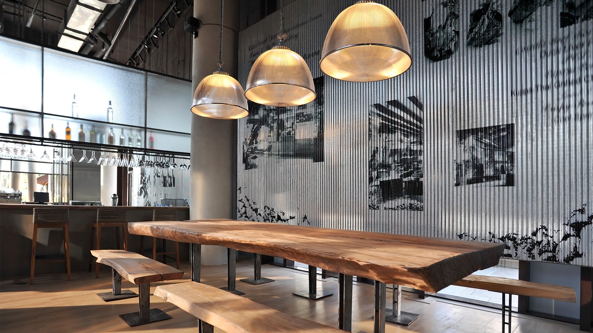 Restaurant interior with a long live-edge communal table, pendant lights, and a bar by large windows.