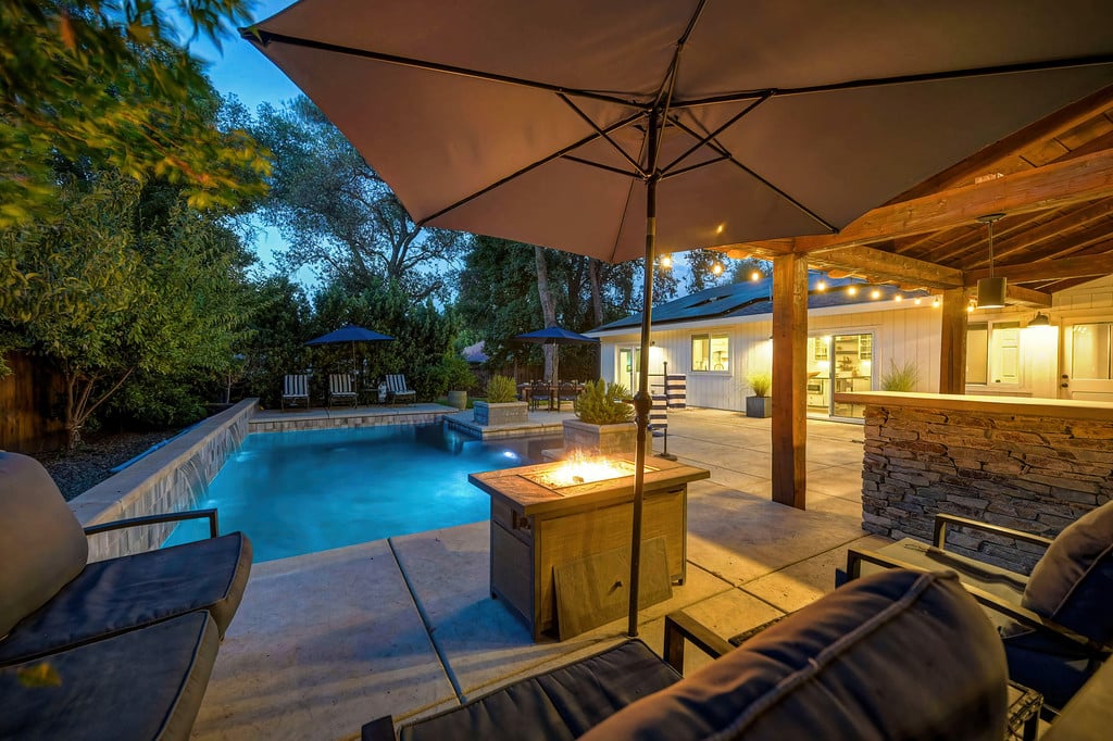 Backyard pool beside a covered patio with cushioned seating and a fire table at dusk.