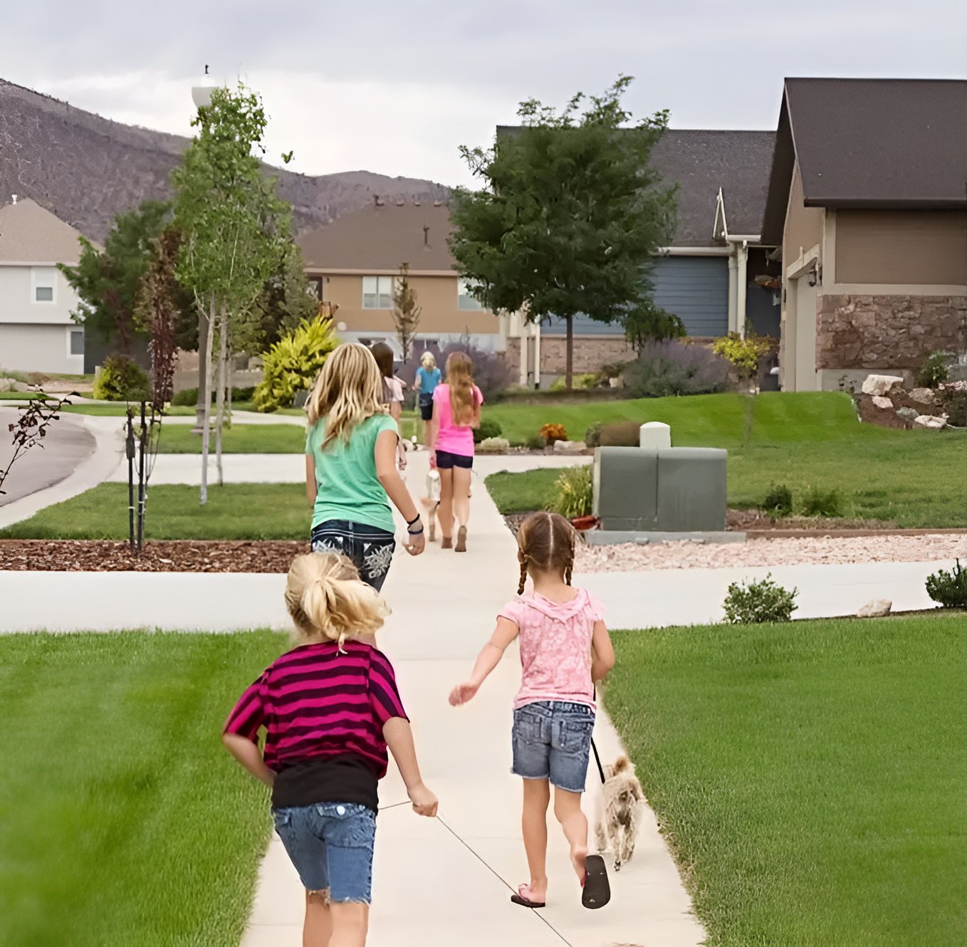 Children and an adult walk a dog along a sidewalk in a landscaped neighborhood.