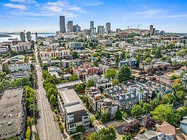 Aerial view of a Seattle neighborhood with downtown skyline, tree-lined streets, and water in the distance.