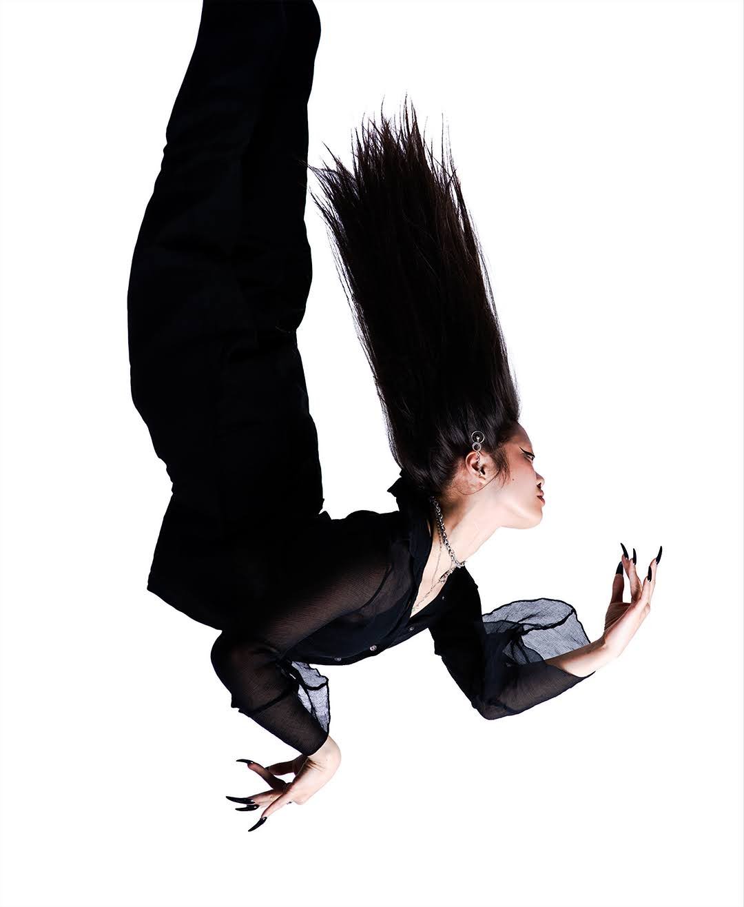 Dancer in black contorts in midair, long hair flying upward against white.
