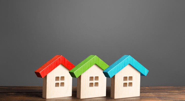 Three small wooden houses with colorful roofs—red, green, and blue—sit on a wooden surface against a gray background, creating a playful and inviting scene.