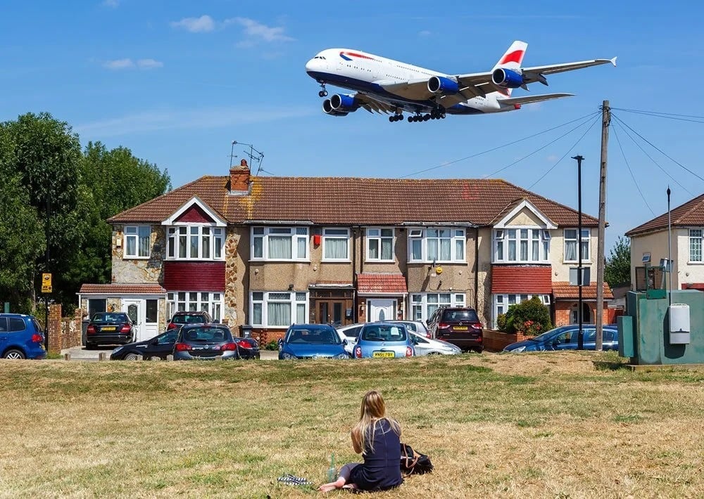 Row of homes beneath a low-flying passenger jet, with a person sitting in the grass.