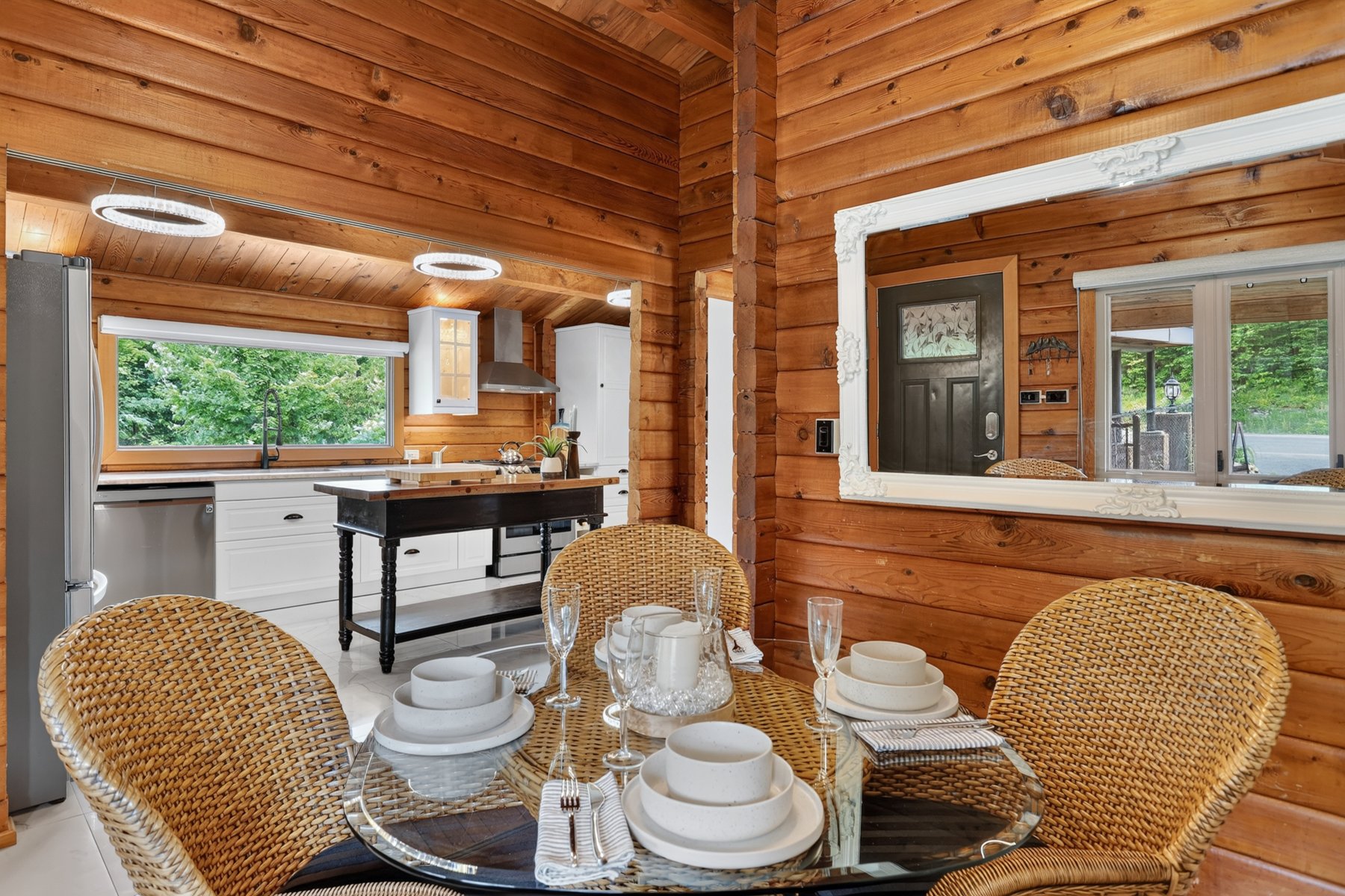 Dining area with a set glass table and wicker chairs beside a white kitchen in a log-walled home.