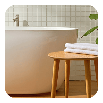 Freestanding bathtub against square tile, with folded towels on a wooden stool.