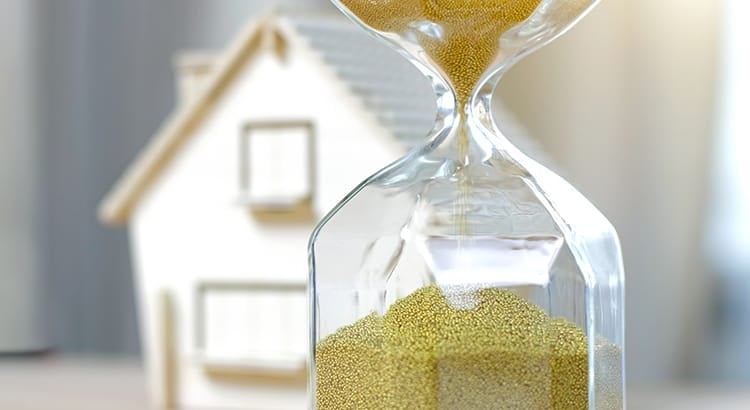 The sand is a golden color, symbolizing the passage of time or urgency. In the background, a blurred-out house model is visible, indicating a potential connection to real estate or home ownership. The focus on the hourglass and the house suggests themes of timing, possibly related to buying or selling a home, real estate investment, or the urgency of decisions in the housing market.
