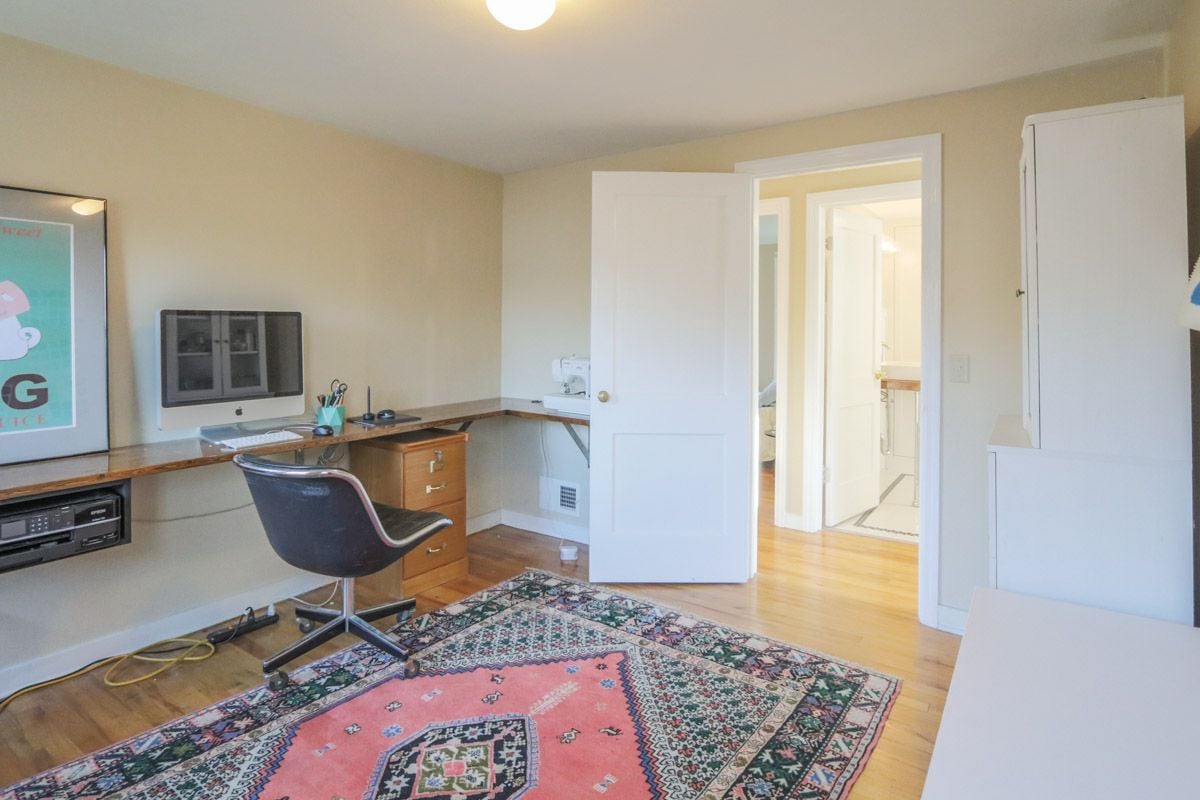 Home office with built-in wooden desk, dark swivel chair, and colorful patterned rug.