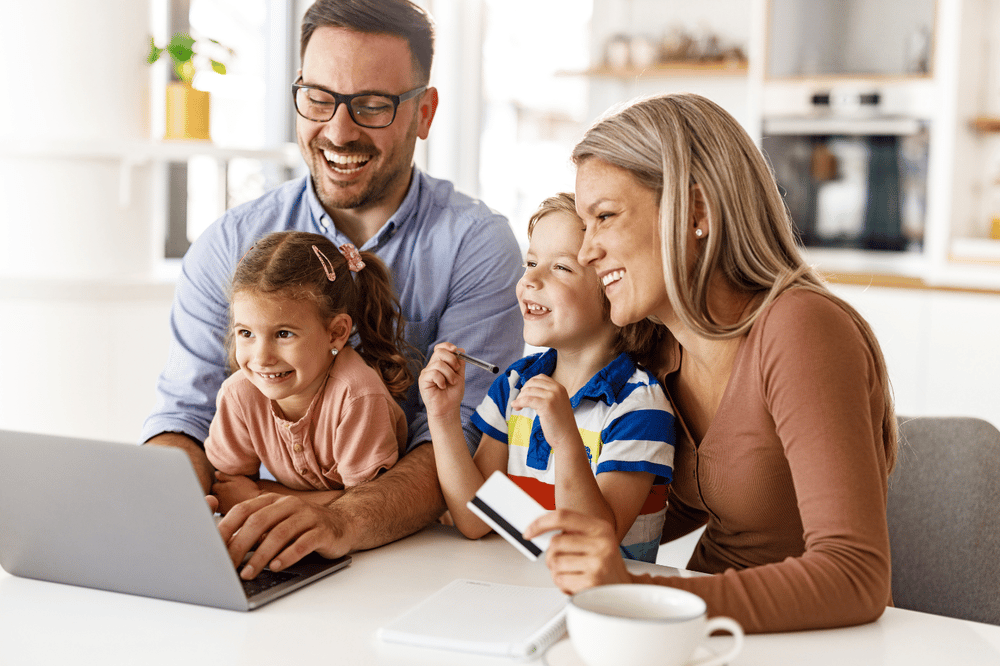 Family gathers around a laptop at the kitchen table, with a child holding a pencil.