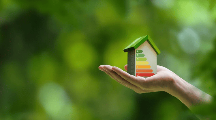 A hand holds a miniature house with a colored energy-efficiency rating scale.