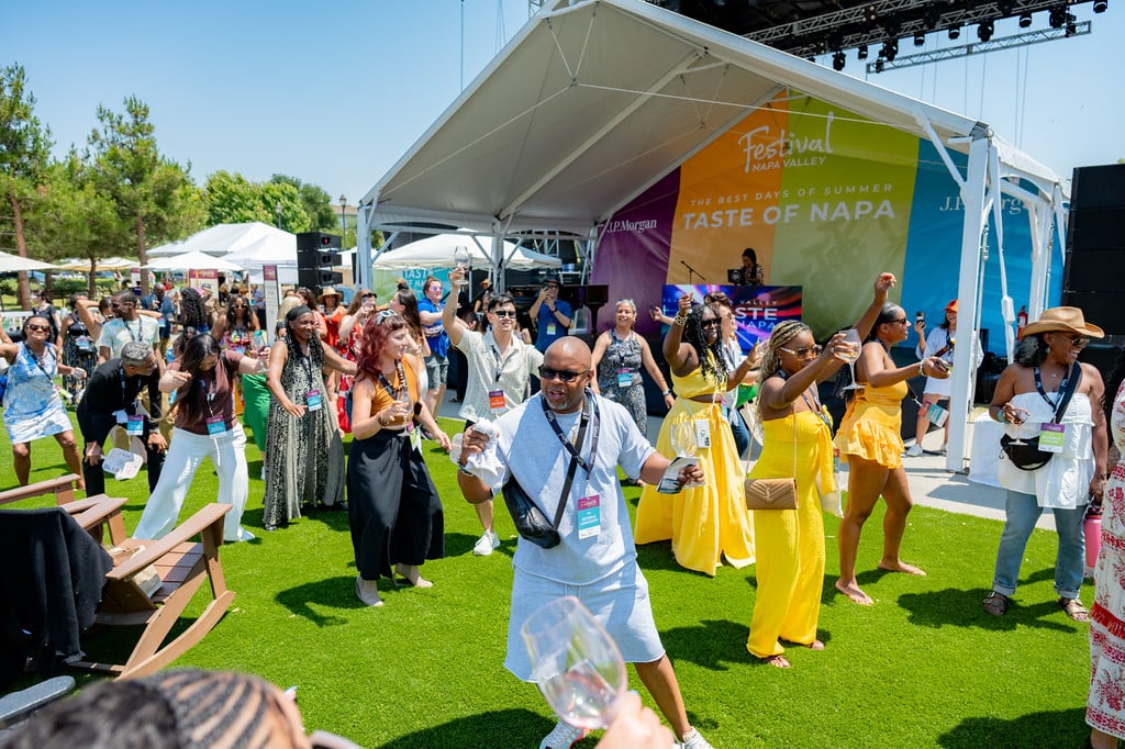 Festival Napa Valley guests dance on a sunny lawn in front of the Taste of Napa stage, holding wine glasses during an outdoor summer event.