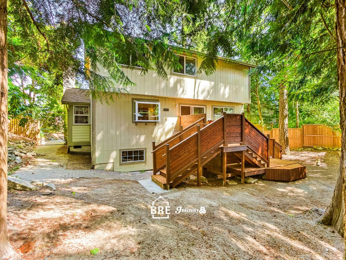 Two-story house with a wooden rear staircase and deck beneath mature trees.