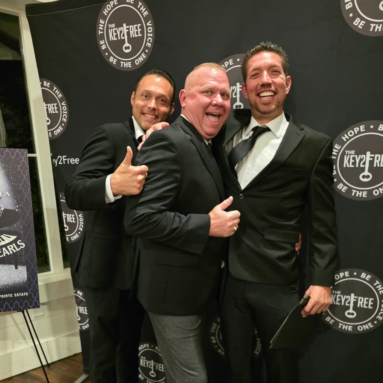 Three men in suits pose in front of a Key2Free backdrop; the man on the left gives a thumbs-up.