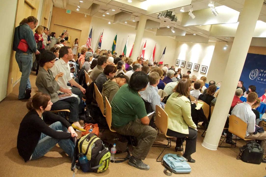 A crowded indoor venue where an audience is closely seated in chairs and on the floor, listening to a panel on the right side of the room, with several international flags displayed behind the panel and a sign for the 
