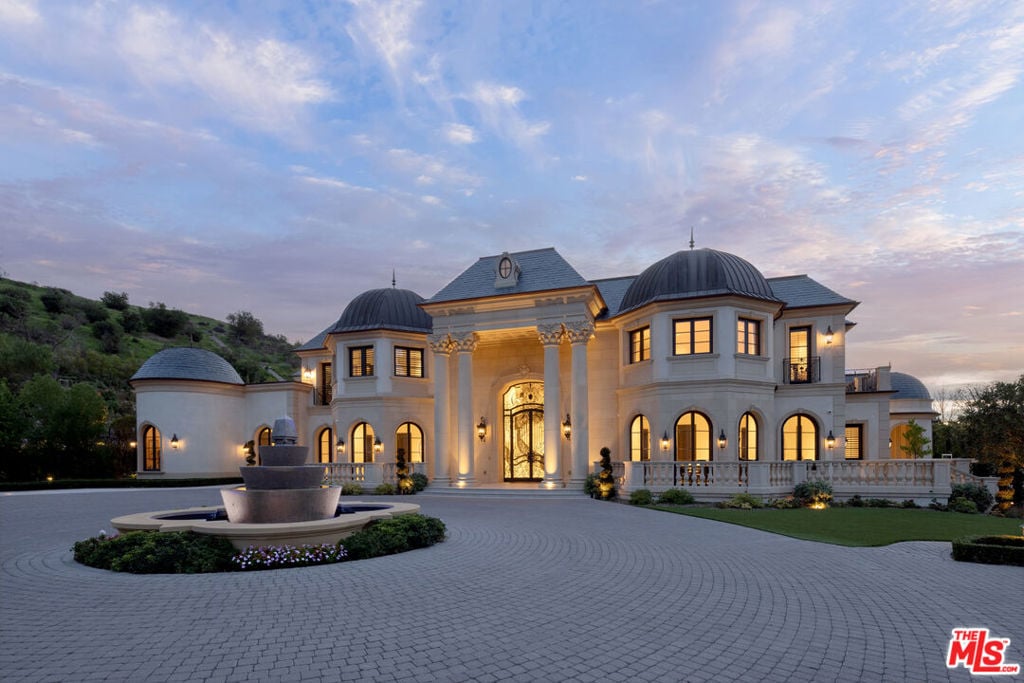 Two-story estate with domed roofs, a circular driveway, and a tiered fountain at dusk.