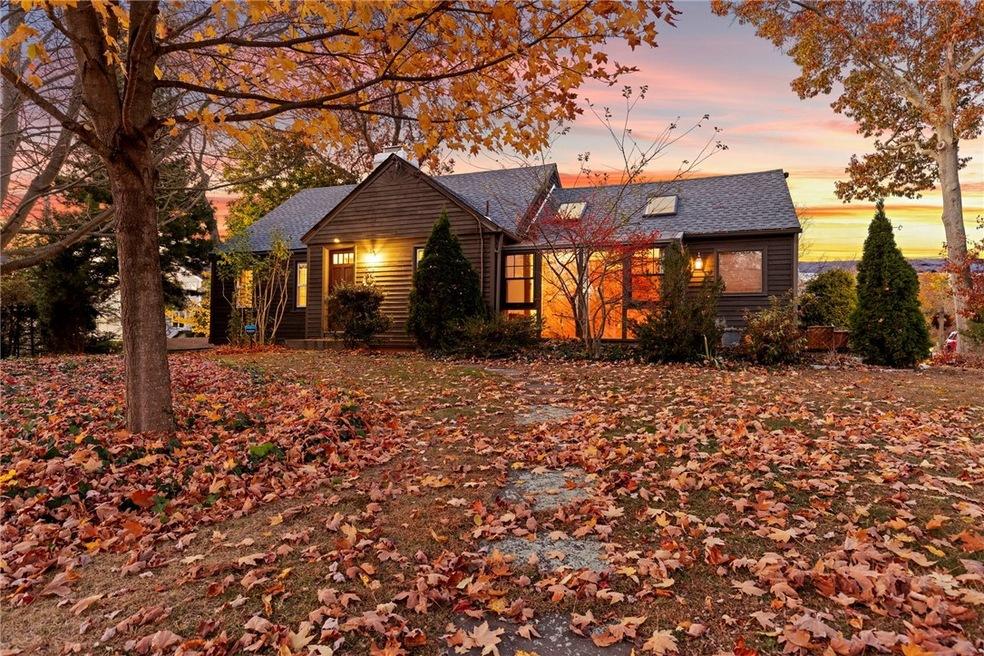 Dark-shingled house glowing at dusk, framed by autumn trees with water visible beyond.