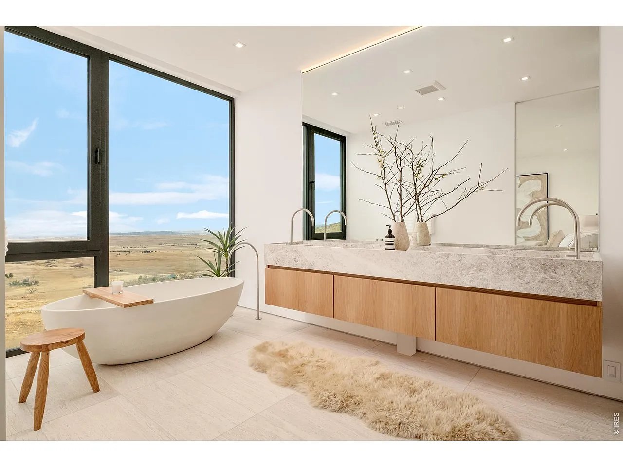 Modern bathroom with a freestanding tub, long wood-fronted vanity, and wide countryside views through large windows.