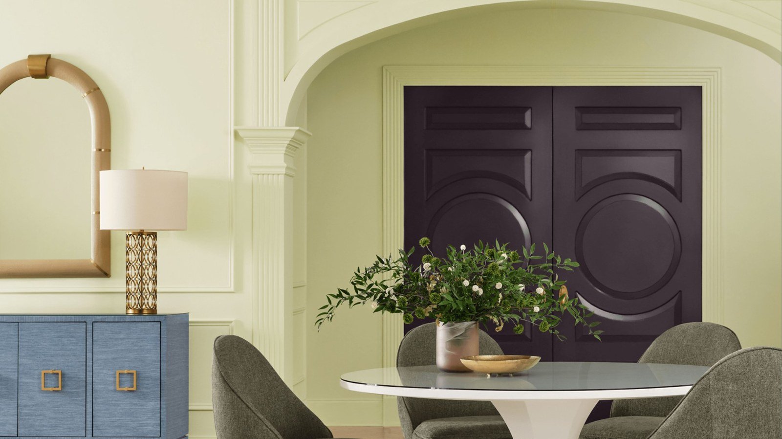 A dining room with walls painted Celery SW 6421, an arched opening, a round white pedestal table with gray chairs, a blue sideboard and a dark paneled door.