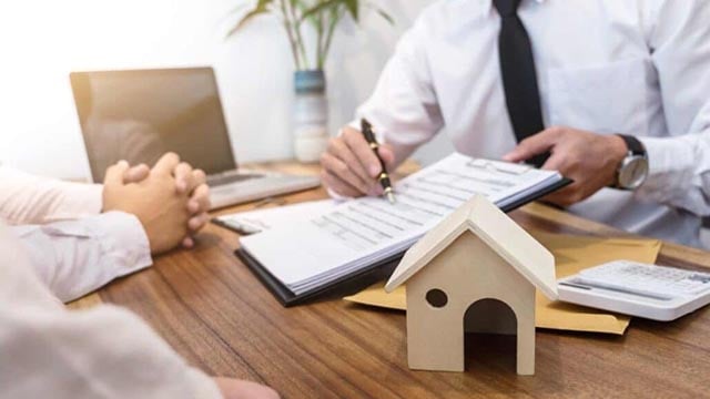 A person reviewing documents with someone at a desk, with a small wooden house model and calculator nearby