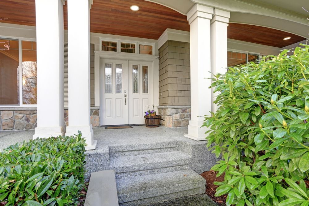 Covered front entry with white double doors, a wood-plank ceiling, stone steps, and leafy shrubs.