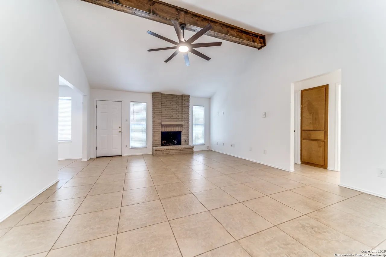 Empty living room with a brick fireplace, large tile floors, and an exposed wood ceiling beam.