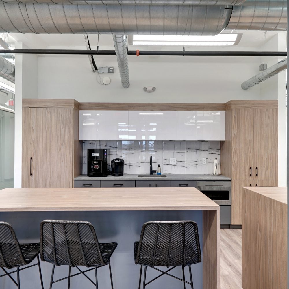 Modern kitchenette with a wood-grain island, black woven stools, and exposed ceiling ductwork.