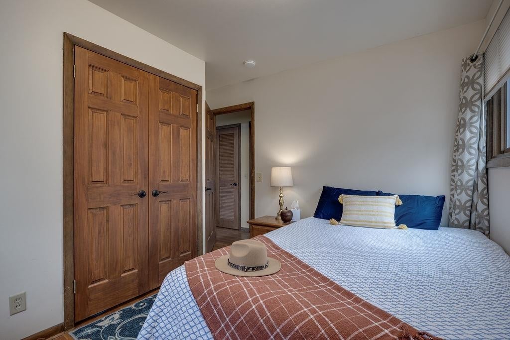 Bedroom with a flat-brimmed hat on a rust-colored throw, blue pillows, and wooden closet doors.