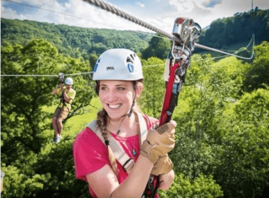 A smiling woman ziplining above a green mountain forest, with another rider in the distance.
