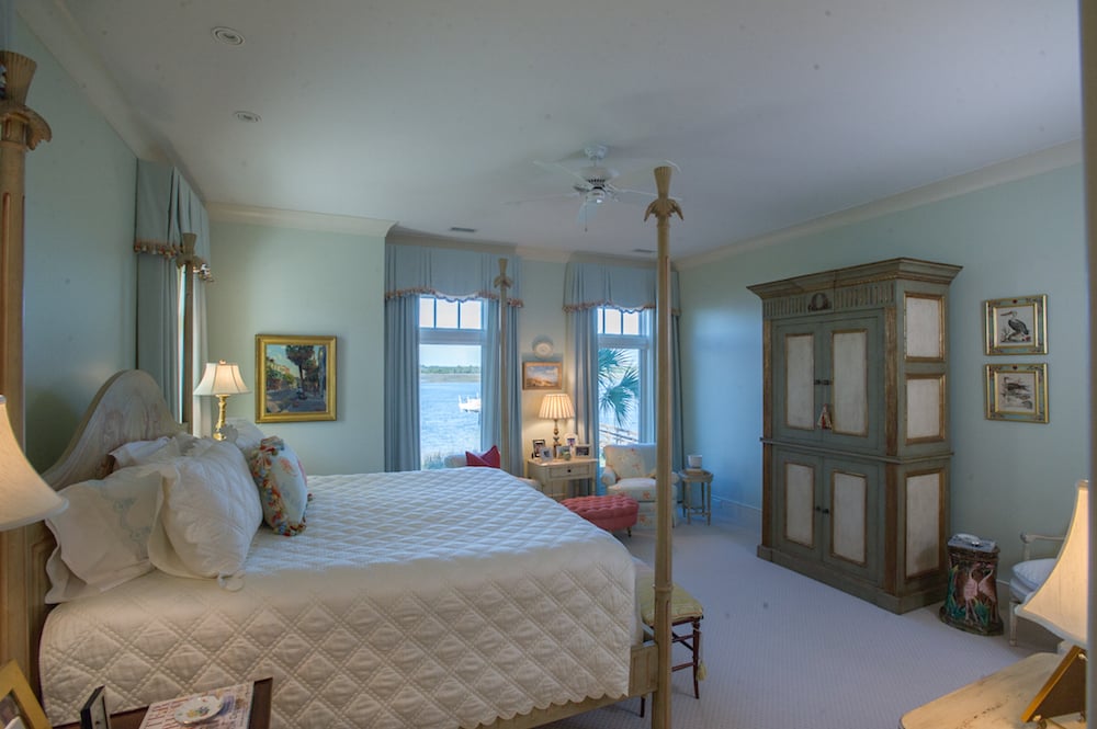 Master bedroom with a large quilted bed, painted armoire, and windows overlooking the water.