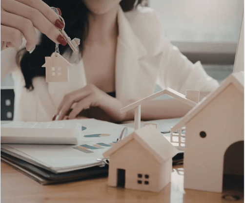 Woman holding a house-shaped keychain above miniature homes and property documents.