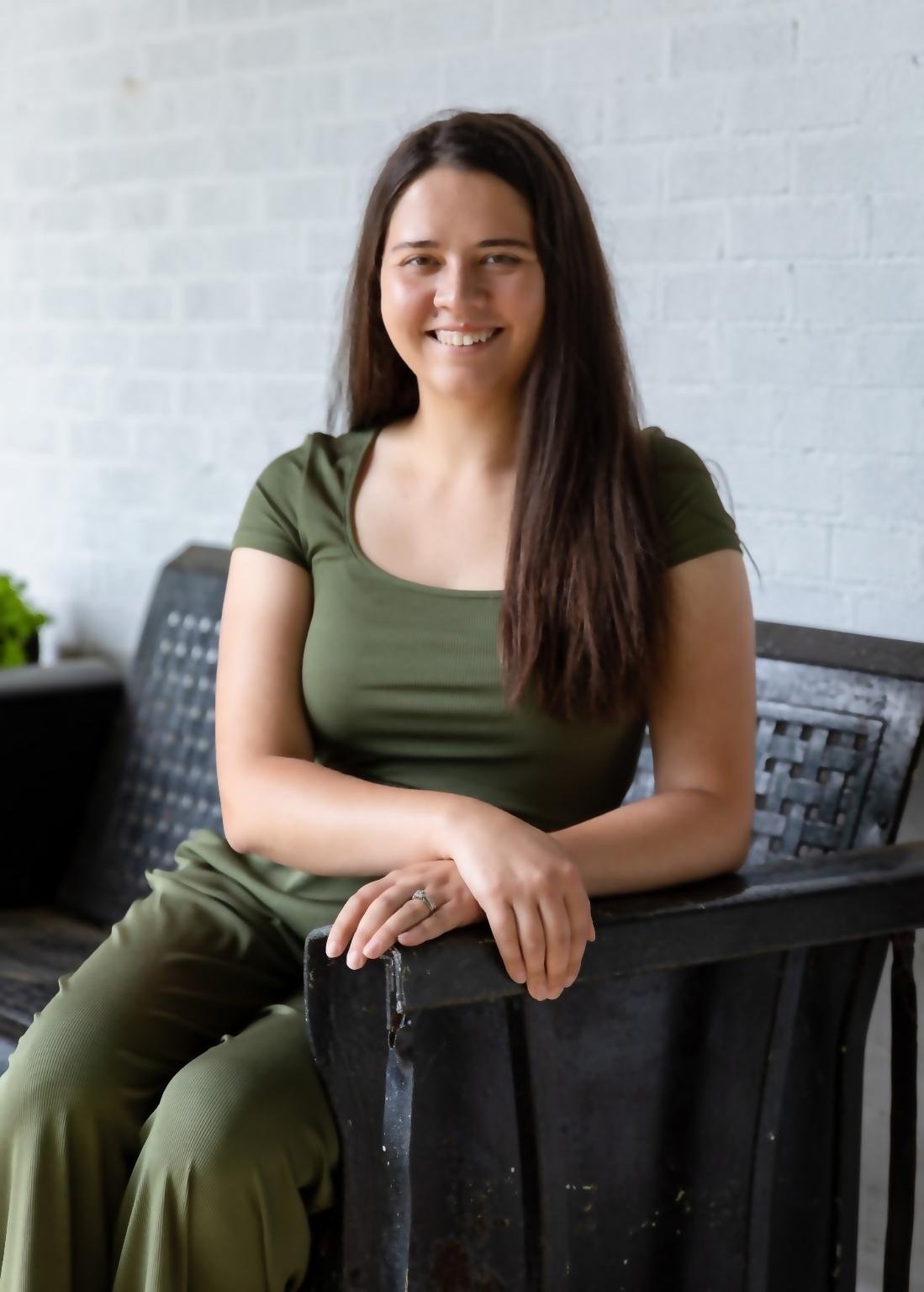 Brittany Gonzalez seated in an olive green outfit, smiling against a white brick wall.