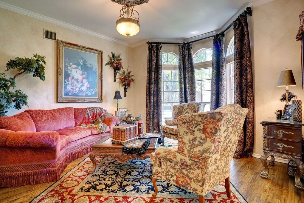 Living room with a rose-pink sofa, floral armchairs, patterned rugs, and tall arched windows.