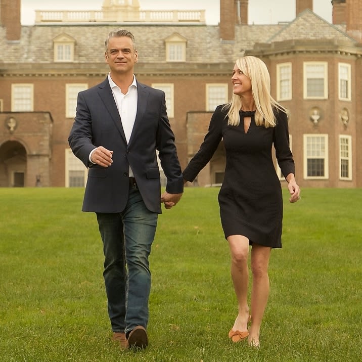  A middle-aged couple walks hand-in-hand across a grassy lawn with a large, stately brick building in the background. 