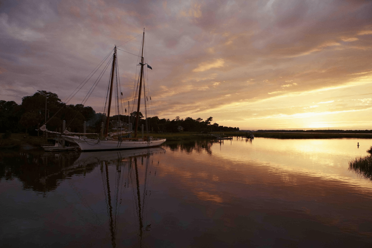 Sailboats reflected in calm marsh water beneath a golden sunset sky.