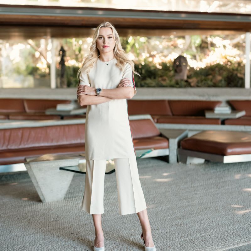 Blonde woman in a white outfit stands with crossed arms in a modern lounge.