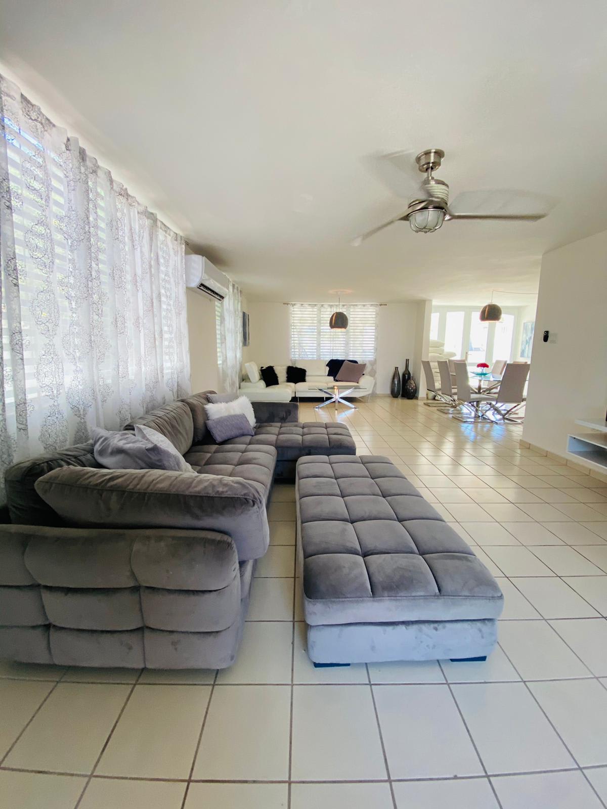 A cozy living room with a couch, ottoman, and ceiling fan. The couch is gray and has a cushion on it.