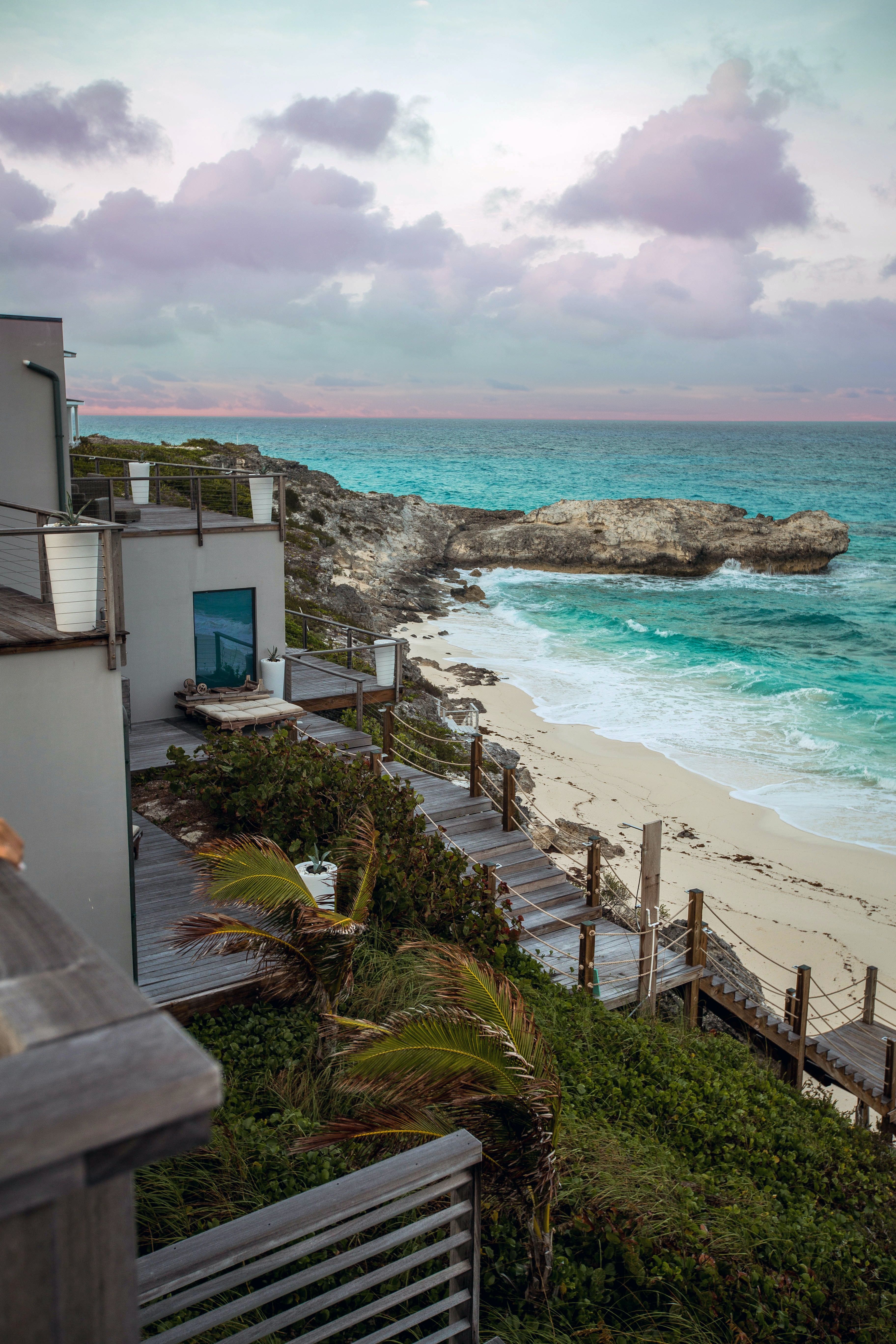 Coastal home with tiered decks overlooking a rocky beach and turquoise ocean at dusk.
