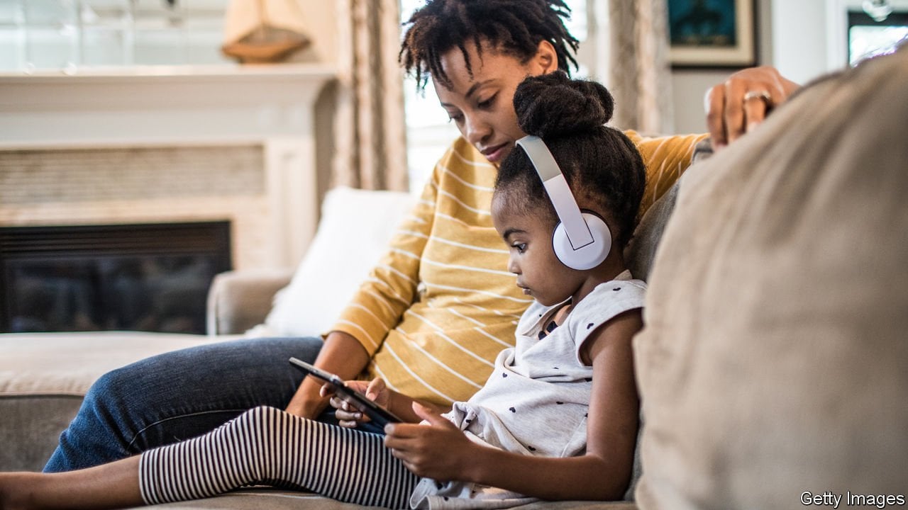 A mother sits with a child wearing headphones and using a tablet on a sofa.