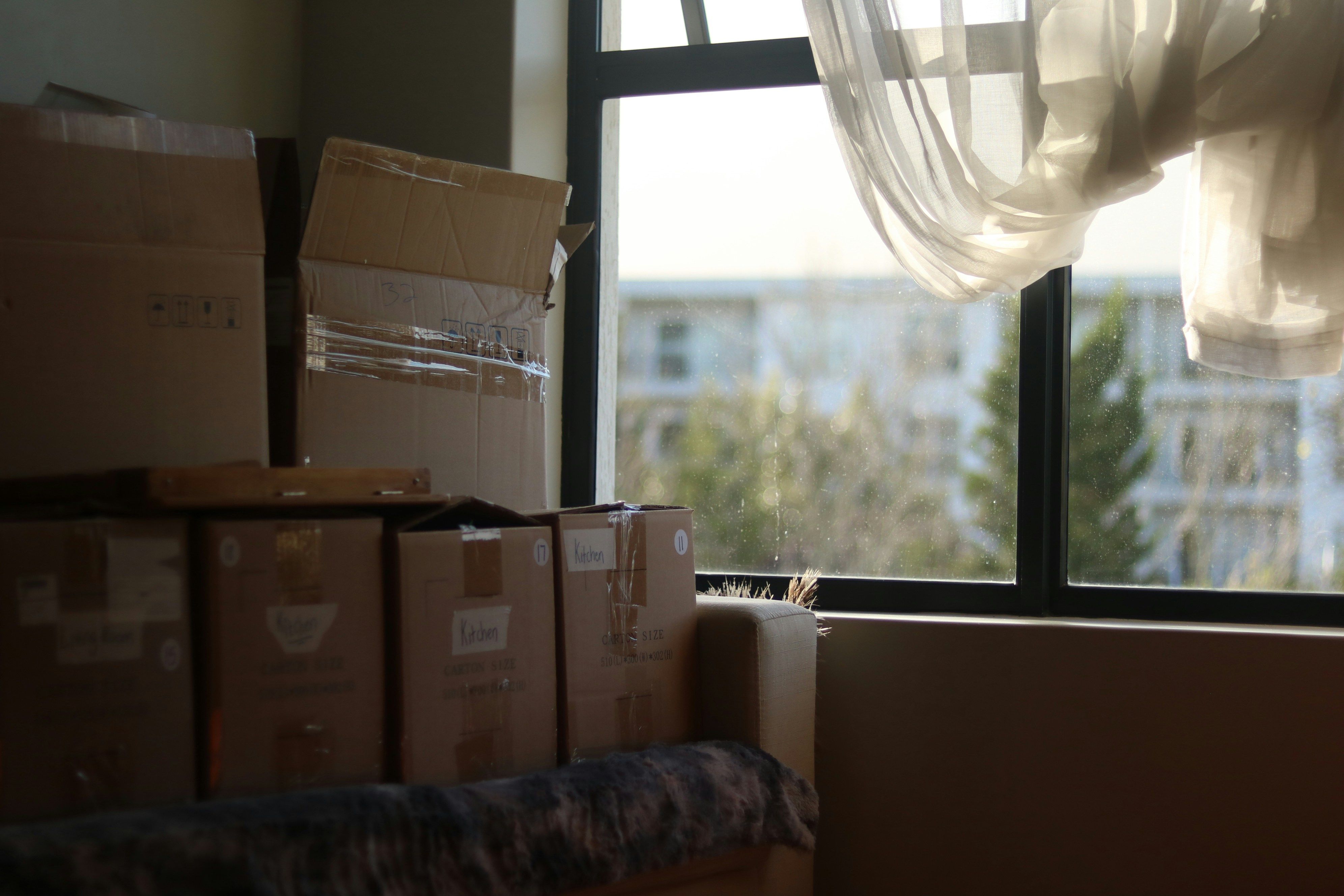 Moving boxes stacked beside a sunlit window with sheer curtains.
