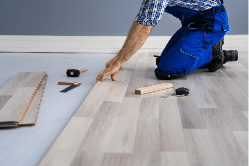 Flooring installer fitting a pale wood-look plank, with spare boards and tools nearby.
