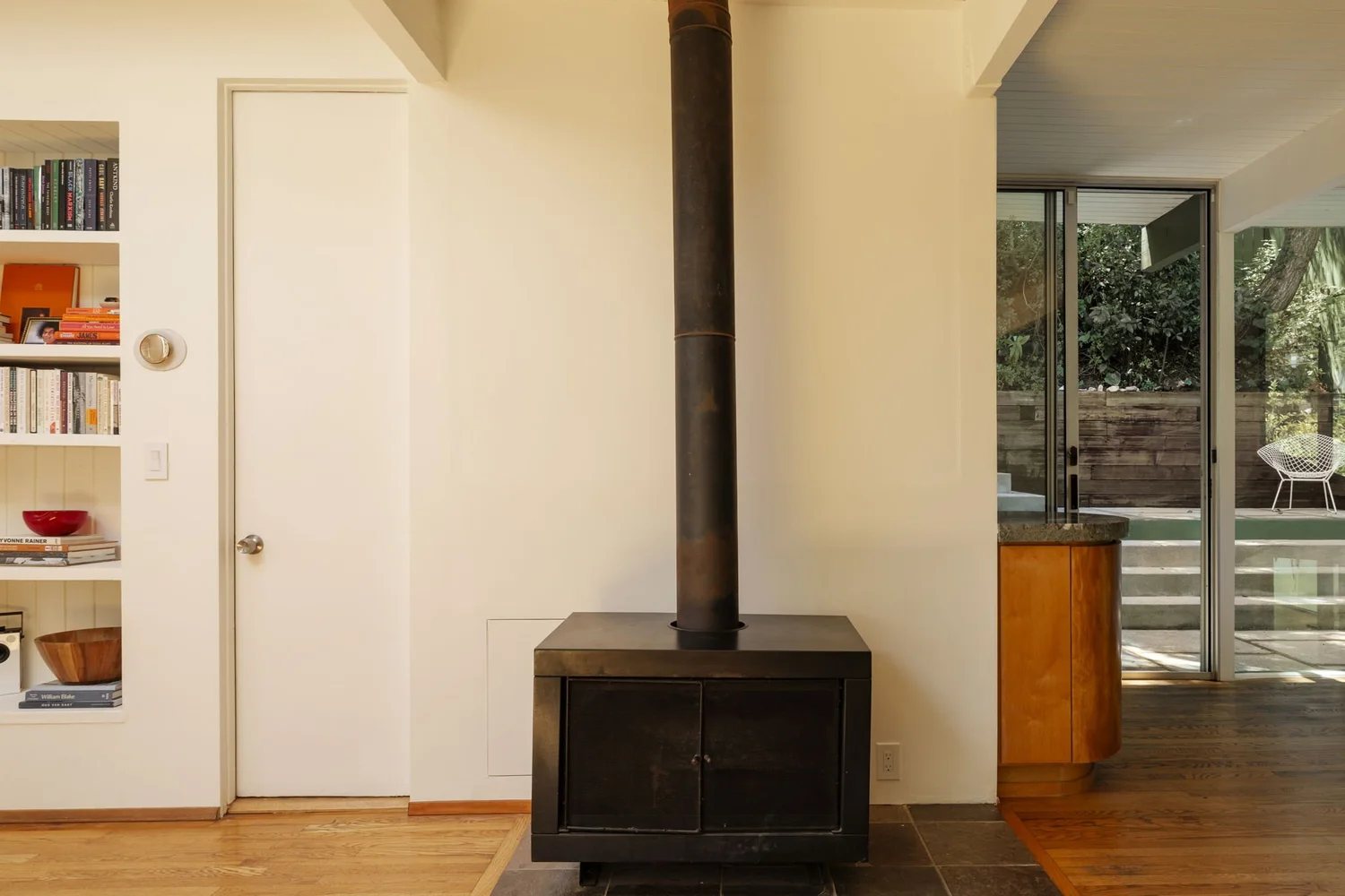 A black freestanding fireplace with a tall metal flue stands on a slate hearth against a white wall, next to built-in bookshelves.
