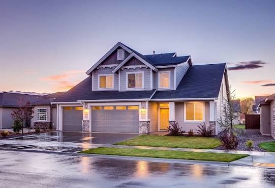 Exterior of a two-story suburban home at dusk with warm interior lighting and a wet street in front